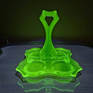 May include: A bright green, vintage glass serving piece with a handle and two circular indentations. The piece has a unique, wavy design and is likely from the Art Deco era. The glass glows under a black light, showcasing its vibrant color.