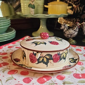 May include: A cream-colored ceramic butter dish with a lid, decorated with red strawberries and green leaves. The dish sits on a matching plate, and the lid has a red strawberry-shaped knob. The design is a vintage style.