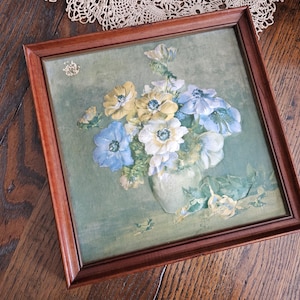 May include: A framed floral painting featuring a bouquet of blue, white, and yellow flowers in a white vase. The artwork is set against a light green background and framed in a dark brown wooden frame. The painting is a still life.