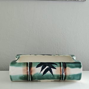 May include: Rectangular ceramic planter with a bamboo design. The planter features a cream-colored interior and a green, brown, and white exterior. Dark green leaf accents are painted on the front. The planter measures approximately 10 inches long.