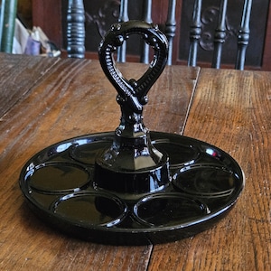 May include: A black, vintage, oval-shaped serving tray with six circular indentations. The tray has a decorative handle and is made of a glossy material. The tray is sitting on a wooden surface.
