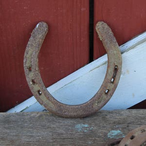 May include: A rusty horseshoe with nail holes, resting on a weathered wooden surface. The horseshoe is positioned against a red and white painted wooden background.