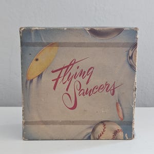 May include: Vintage box with a brown background and red text that reads "Flying Saucers". The box has illustrations of flying saucers and baseballs.