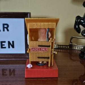 May include: A novelty figurine of a wooden outhouse with a figure inside. The outhouse is tan with a red base and a sign that reads "LADIES ONLY". A sign that reads "BAR OPEN" is in the background.