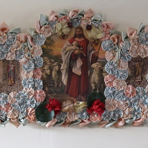 May include: A framed religious image of Jesus as the Good Shepherd, surrounded by a wreath of fabric flowers in shades of pink and blue. The image depicts Jesus holding a lamb in his arms, with a flock of sheep in the background.