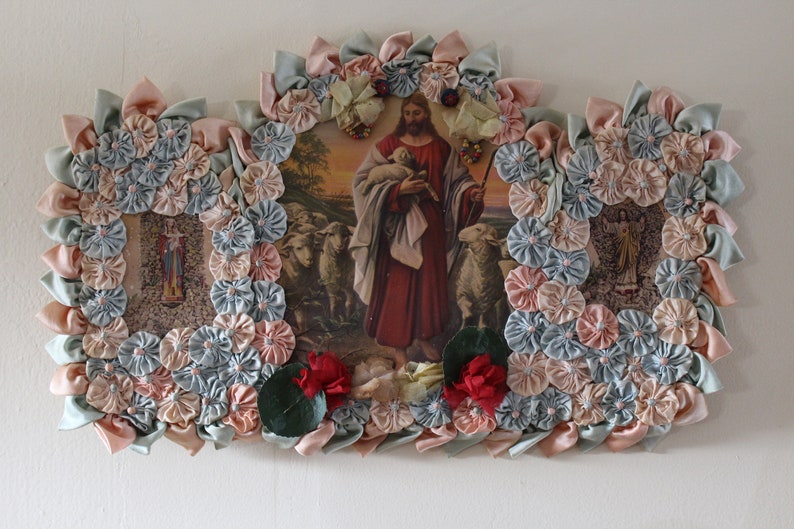May include: A framed religious image of Jesus as the Good Shepherd, surrounded by a wreath of fabric flowers in shades of pink and blue. The image depicts Jesus holding a lamb in his arms, with a flock of sheep in the background.