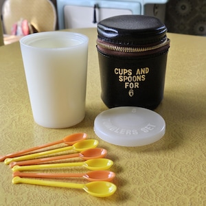 May include: A black leather travel case with gold lettering that reads "Cups and Spoons for 6" and a white plastic container with a white lid. The case is open and the container is sitting on a yellow surface. There are six yellow and orange plastic spoons on the surface.