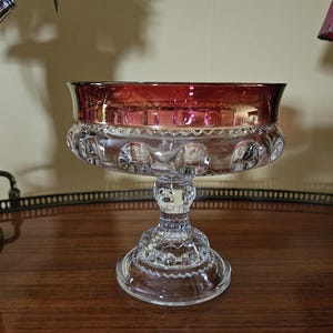 May include: A clear glass footed bowl with a ruby red rim. The bowl has a decorative pattern around the base and the bowl's rim. The bowl is sitting on a wooden surface.