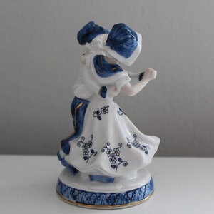 May include: A porcelain figurine of a couple dancing, painted in blue and white with gold accents. The woman wears a white dress with blue floral designs and a blue hat. The man wears a blue and white patterned outfit. The figurine is on a blue and white base.