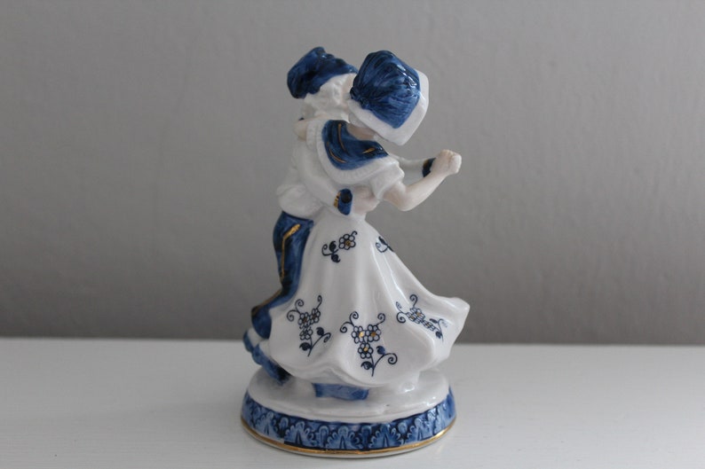 May include: A porcelain figurine of a couple dancing, painted in blue and white with gold accents. The woman wears a white dress with blue floral designs and a blue hat. The man wears a blue and white patterned outfit. The figurine is on a blue and white base.