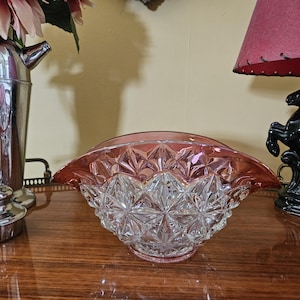 May include: A clear glass bowl with a pink rim and a starburst pattern. The bowl is sitting on a wooden surface. A silver cocktail shaker and a black horse lamp are in the background.