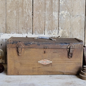 May include: A rectangular brown metal Craftsman toolbox with a top handle and two front latches. The box features a central oval logo with the text Craftsman. To the left is a glass oil lamp, and to the right are three stacked weights.