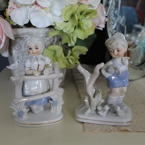 May include: Two porcelain figurines featuring a young person in a white bonnet and blue skirt leaning on a fence, and a young person in a blue cap and blue shorts standing near a tree stump with two small birds at their feet, both with gold trim.