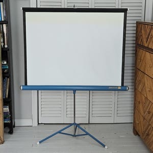 May include: A portable Da-Lite projection screen featuring a rectangular white surface framed by a black border. The screen is mounted on a blue metal tripod stand with a central vertical pole and three legs extending outward for stability.