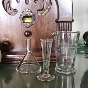 May include: Three clear glass laboratory items: a funnel, a small vase, and a graduated beaker with the text "Eastman Kodak Rochester". A decorative black crow figurine is on the right. The background includes a vintage wooden radio.