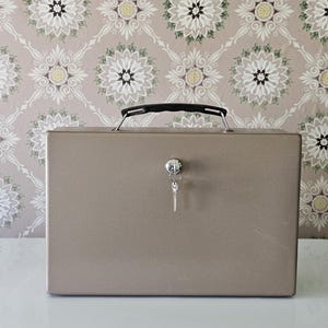 May include: A rectangular, metallic beige lockbox featuring a black plastic carrying handle on top. A silver-toned circular keyhole is centered on the front panel, with a small metal key inserted and hanging downwards from the lock mechanism.