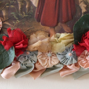 May include: A vintage picture frame decorated with fabric flowers in shades of pink, blue, and red. The flowers are arranged around the frame, creating a border. There are also green leaves and a piece of yellow fabric.