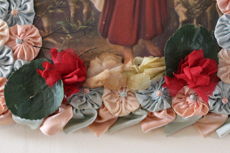 May include: A vintage picture frame decorated with fabric flowers in shades of pink, blue, and red. The flowers are arranged around the frame, creating a border. There are also green leaves and a piece of yellow fabric.