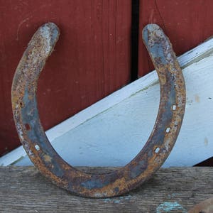 May include: A rusty horseshoe with a worn, weathered surface. The horseshoe is lying on a wooden surface with a white and red painted background.