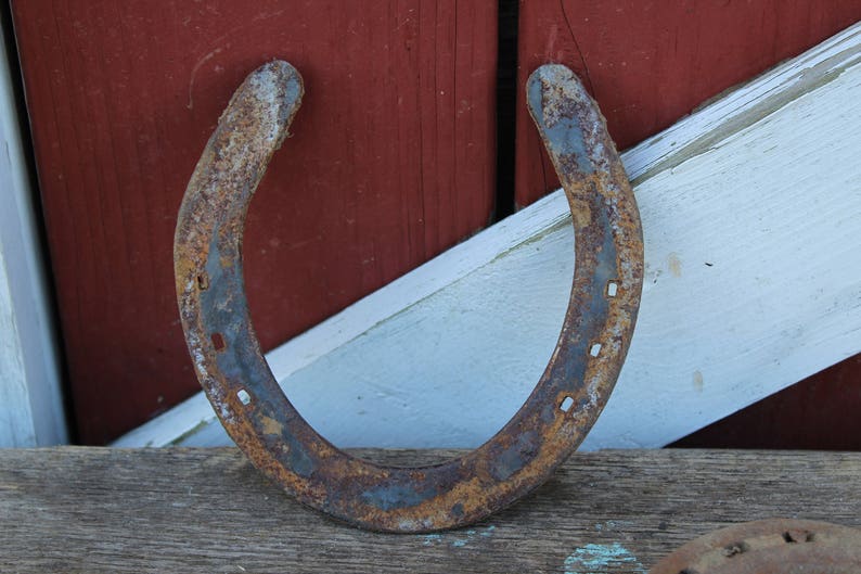 May include: A rusty horseshoe with a worn, weathered surface. The horseshoe is lying on a wooden surface with a white and red painted background.
