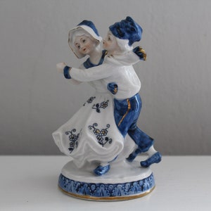 May include: A porcelain figurine of a couple dancing. The woman is wearing a white dress with blue and gold accents. The man is wearing blue trousers and a blue and white shirt. The figurine is on a white base with blue and gold accents.