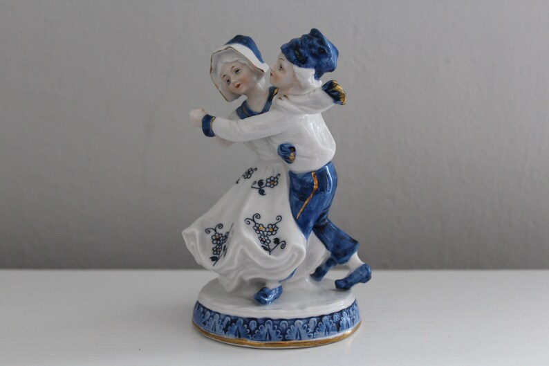 May include: A porcelain figurine of a couple dancing. The woman is wearing a white dress with blue and gold accents. The man is wearing blue trousers and a blue and white shirt. The figurine is on a white base with blue and gold accents.