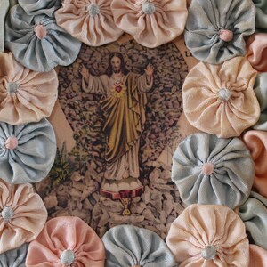 May include: A framed image of Jesus Christ with a heart on his chest, surrounded by a wreath of light blue and peach fabric flowers.
