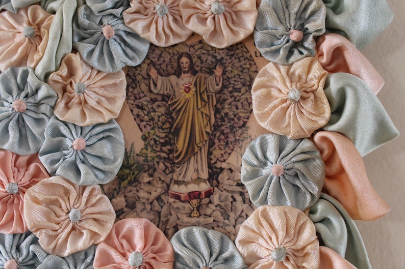 May include: A framed image of Jesus Christ with a heart on his chest, surrounded by a wreath of light blue and peach fabric flowers.