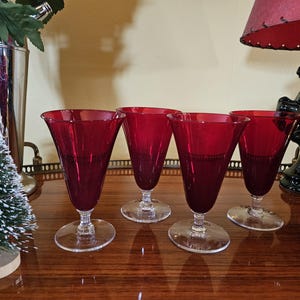 May include: Four clear glass stemmed wine glasses with a deep red hue. The glasses are arranged in a row on a wooden surface.