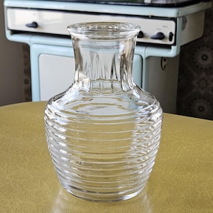 May include: A clear glass decanter with a ribbed, horizontal design on the lower portion and vertical ridges on the neck. The decanter sits on a yellow surface, with a vintage stove visible in the background.