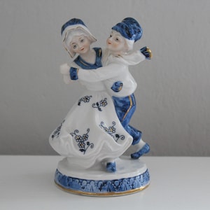 May include: A porcelain figurine of a couple dancing. The woman is wearing a white dress with blue and gold floral designs. The man is wearing a blue and white outfit with a blue hat. The figurine is on a blue and gold base.