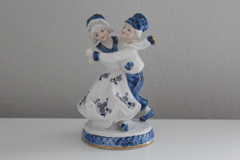 May include: A porcelain figurine of a couple dancing. The woman is wearing a white dress with blue and gold floral designs. The man is wearing a blue and white outfit with a blue hat. The figurine is on a blue and gold base.