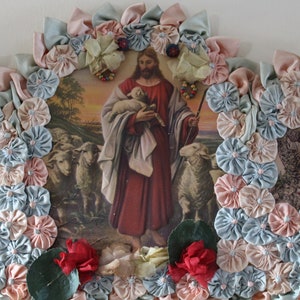 May include: A framed religious image of Jesus holding a lamb with a flock of sheep in the background. The image is surrounded by a border of blue and pink fabric rosettes.