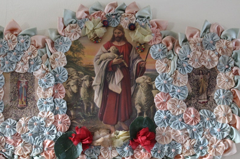 May include: A framed religious image of Jesus holding a lamb with a flock of sheep in the background. The image is surrounded by a border of blue and pink fabric rosettes.