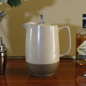 May include: A two-toned ceramic pitcher featuring a cream-colored upper body and a taupe-colored base. The pitcher includes a matching lid topped with a silver-toned metal finial, a curved handle, and a wide pouring spout for liquid service.