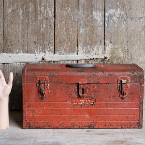 May include: A rectangular red metal toolbox with a weathered, distressed finish, featuring two front latches, a central locking mechanism, and a black handle on top. The box has horizontal embossed lines and a small, worn logo plate on the front.