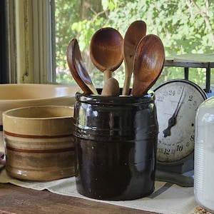 May include: A dark brown cylindrical ceramic crock filled with several wooden spoons, positioned next to a tan bowl with brown stripes, a light green bowl, a vintage metal scale with a round dial, and a clear glass jar containing white powder.