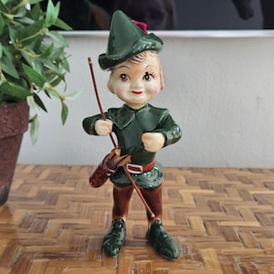 May include: A ceramic figurine of a young boy dressed in a green outfit with a matching hat and brown boots. The figurine is holding a bow and arrow and has a quiver on its side. The boy has rosy cheeks and is standing on a wooden surface.