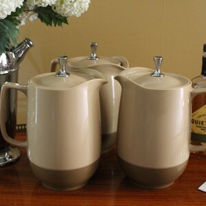 May include: Three beige thermal carafes with silver lids and handles on a wooden surface. The carafes are in a row, with the middle carafe slightly behind the other two. There are other items on the surface, including a silver cocktail shaker, a silver cocktail shaker, and two bottles of liquor.