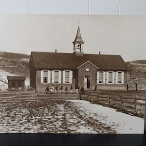 May include: A black and white photograph of a brick schoolhouse with a bell tower. The schoolhouse has a wooden fence in front of it and a small outhouse to the left. There are children standing in front of the schoolhouse.