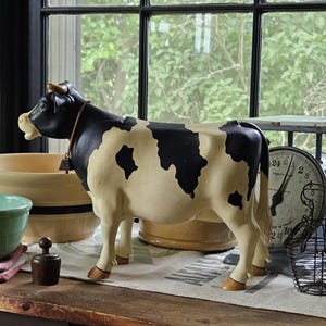 May include: A black and white cow figurine with a small bell on a thin cord around its neck. The cow is standing in profile, featuring a cream-colored body with irregular black patches, a dark head, and small horns, positioned among various kitchen items.