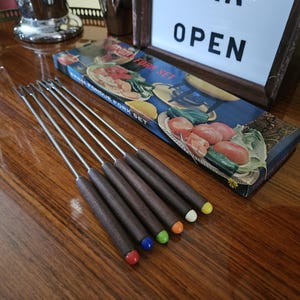 May include: A set of six stainless steel fondue forks with long, dark brown wooden handles, each featuring a distinct colored tip in red, blue, green, orange, white, and yellow. The forks are displayed next to their original rectangular box.