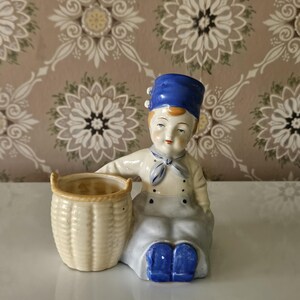 May include: A ceramic figurine of a seated chef with a blue hat and shoes, white uniform, and a basket. The figurine is painted in soft colors and has a detailed design. The basket is beige and has a woven texture. The background is a floral wallpaper.