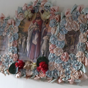 May include: A framed religious image of Jesus holding a lamb surrounded by blue and pink fabric flowers. The image is framed with a decorative border of fabric flowers.