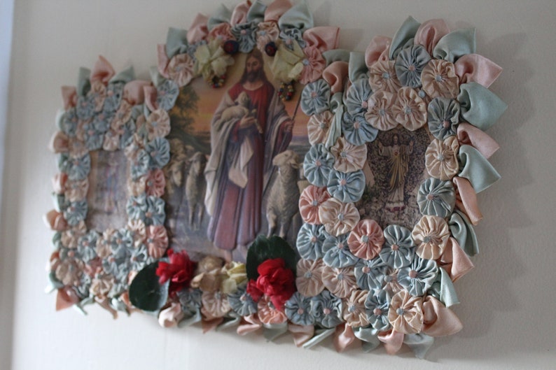 May include: A framed religious image of Jesus holding a lamb surrounded by blue and pink fabric flowers. The image is framed with a decorative border of fabric flowers.