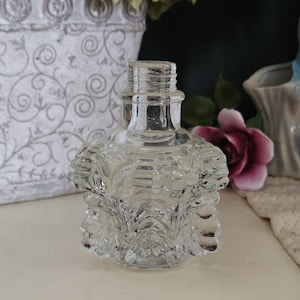 May include: A clear, vintage-style glass perfume bottle with a textured, ornate design. The bottle has a fluted neck and a decorative, ruffled body. The background includes a white floral patterned container, a pink rose, and a ceramic bird figurine.