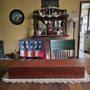 May include: A rectangular wooden box with a dark reddish-brown finish, featuring a flat lid and a smooth, solid construction. The box is positioned horizontally on a white lace doily, showcasing a simple, functional design with visible wood grain.