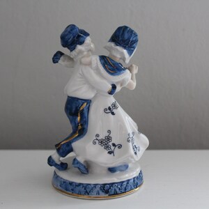 May include: A porcelain figurine of a couple in traditional Dutch clothing, dancing. The figurine is painted in blue and white with gold accents.