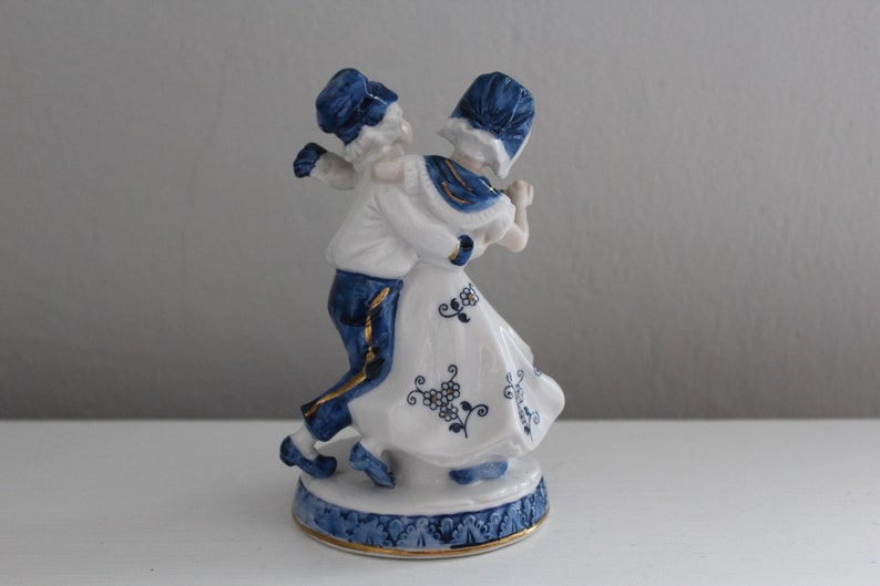 May include: A porcelain figurine of a couple in traditional Dutch clothing, dancing. The figurine is painted in blue and white with gold accents.