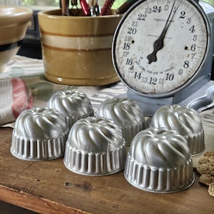 May include: A set of six individual silver-colored metal fluted molds with a circular, spiraled design. Each mold features a ridged side and a smooth, rounded top, arranged in a cluster on a wooden surface near a vintage kitchen scale.